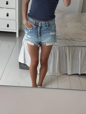 Blå denimshorts med fransig kant - Säljer ett par ljusblå denimshorts med hög midja och fransig kant. Shortsen har slitna detaljer och klassiska fickor både fram och bak. Perfekta för sommaren och ger en avslappnad look.