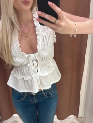 Vit blus med volanger och snörning - Supersöt vit blus med volanger och djup v-ringning. Blusen har snörning framtill och är lätt rynkad för en feminin look. Perfekt att styla med jeans eller kjol för en trendig outfit.