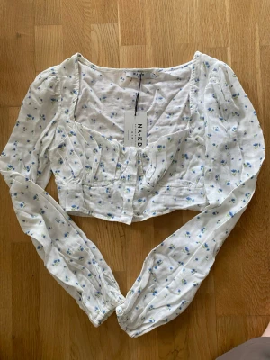 Vit blommig blus från NA-KD - Supersöt vit blus från NA-KD med små blå blommor, lång ärm och knappar framtill. Croppad modell med markerad midja. Perfekt för en trendig och romantisk look.