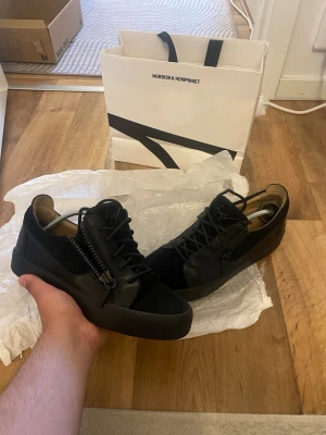 Svarta sneakers från Giuseppe Zanotti - Snygga svarta sneakers från Giuseppe Zanotti med låg profil, dragkedjor på sidorna och klassiska svarta skosnören. Skorna har en stilren design i läder och mocka med diskret logga på plösen. Perfekta för dig som gillar exklusiva detaljer och streetwear-vibe.