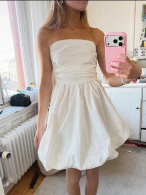Vit axelbandslös kort klänning - Snygg vit axelbandslös kort klänning med ballongformad kjol och markerad midja. Perfekt för dig som vill ha en stilren och fräsch look. Klänningen har en enkel design utan mönster och är tillverkad i ett lätt material. Passar perfekt till student o skolavslutning 