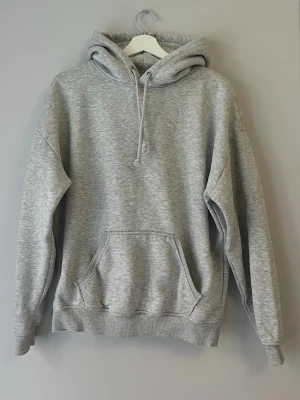 Grå hoodie från BikBok - Klassisk grå hoodie från BikBok med huva och dragsko, samt en stor känguruficka framtill. Mjuk insida och ribbade muddar vid ärmslut och nederkant. Perfekt för en chill och avslappnad stil.