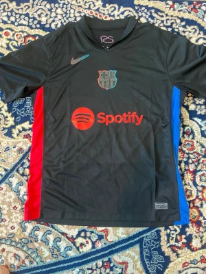 Svart FC Barcelona matchtröja Nike - Säljer en svart FC Barcelona matchtröja från Nike med Dri-FIT material. Tröjan har klubbens emblem på bröstet, Nike-logga och Spotify som sponsor. Sidorna har röda och blå paneler. Perfekt för dig som vill representera Barça på planen.