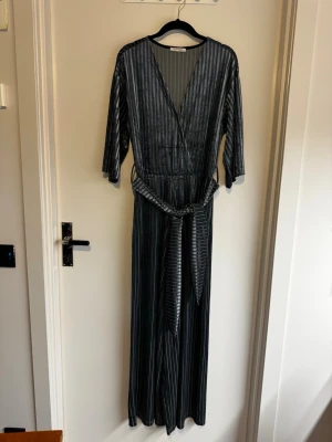Randig jumpsuit från Zara Trafaluc - Snygg svart och grå randig jumpsuit från Zara Trafaluc med omlott-ringning, trekvartsärmar och knytskärp i midjan. Långa, vida ben och mjukt, följsamt plysch- eller velour-material som ger en avslappnad men trendig look.