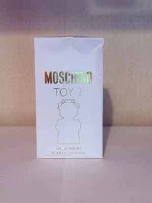 Moschino Toy 2 Eau de Parfum 100 ml - Moschino Toy 2 Eau de Parfum i en unik flaska formad som en nallebjörn. Flaskan är genomskinlig med frostad finish på detaljerna och har ett modernt, lekfullt uttryck. Förpackningen är vit med guldfärgad text och illustration. Volymen är 100 ml.