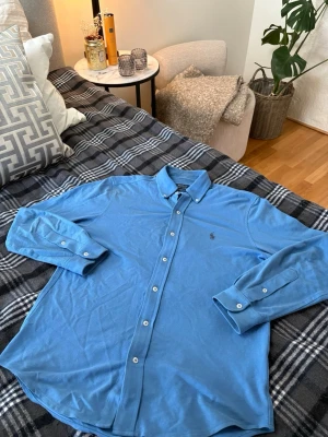 Polo Ralph Lauren Skjorta - Snygg blå featherweight mesh skjorta från Polo Ralph Lauren med klassisk button-down krage och broderad logga på bröstet. Skjortan har långa ärmar och vita knappar framtill. Perfekt för en stilren och avslappnad look.