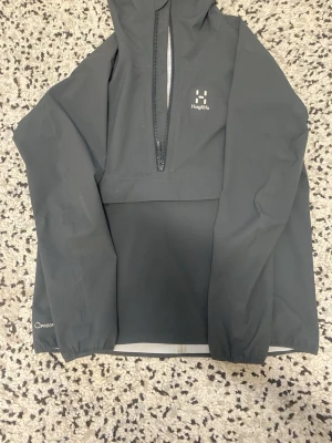 Svart regnjacka från Haglöfs - Snygg svart regnjacka från Haglöfs med halv dragkedja framtill och huva. Jackan har minimalistisk design, diskret logga på bröstet och nederkanten, samt Q Proof-tryck på ärmen. Perfekt för regniga dagar och outdoor-äventyr.