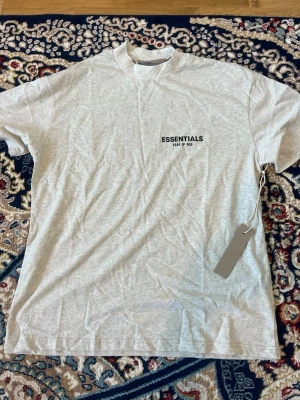 Grå Essentials T-shirt från Fear of God - Snygg grå T-shirt från Fear of God Essentials med svart logga på bröstet. Klassisk passform med rund hals och korta ärmar. Perfekt för en clean och stilren look. Tillverkad i mjuk bomull för maximal komfort.