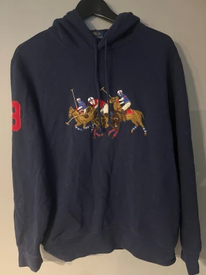 Ralph lauren Hoodie  - Mörkblå hoodie med broderat polospel-motiv på bröstet och röd siffra 3 på ärmen. Klassisk huva med dragsko och mjukt material. Perfekt för en sportig och avslappnad stil.