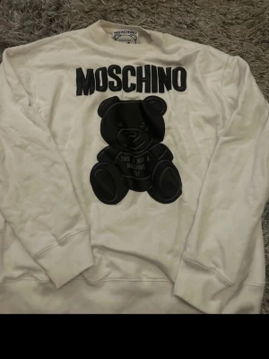 Vit Moschino sweatshirt med björn - Säljer en vit sweatshirt från Moschino med stor svart björn och texten 'This is not a Moschino toy' på framsidan. Tröjan har rund halsringning, långa ärmar och ribbade muddar. Perfekt statement-plagg för dig som gillar streetstyle och designer.