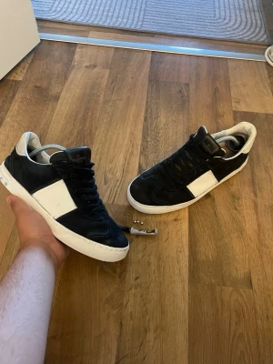 Svarta och vita sneakers från Valentino - Snygga sneakers från Valentino i svart mocka och vitt läder. Skorna har en vit panel på sidan, svarta skosnören och en vit sula med små nitar baktill. Perfekta för dig som gillar stilrena och exklusiva sneakers med en unik touch.