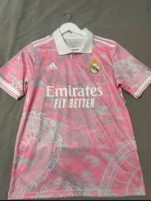 Rosa Real Madrid tröja Adidas - Snygg rosa Real Madrid matchtröja från Adidas med drakmönster i ljusgrått. Tröjan har vit krage, klubbmärke på bröstet och tryck med 'Emirates Fly Better'. Perfekt för fotboll eller streetwear.