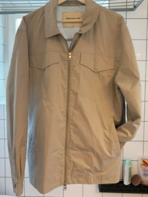 Ljusgrå overshirt från These Glory Days XL (Som Large) - Stilren ljusgrå overshirt med dragkedja framtill från These Glory days. Modellen har klassisk krage, två bröstfickor med lock och knäppning vid ärmslut. Tillverkad i ett lätt och mjukt bomullstyg, perfekt för lager-på-lager.