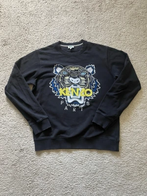 Svart Kenzo sweatshirt med tigertryck - Cool svart sweatshirt från Kenzo med ikoniskt broderat tigerhuvud och texten 'KENZO PARIS' i gult och vitt framtill. Tröjan har rund hals, långa ärmar och ribbade muddar. Perfekt för dig som gillar streetwear och vill sticka ut.