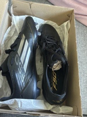 Adidas F50 Pro FG fotbollsskor svart storlek 44  - Säljer ett par svarta Adidas F50 Pro FG fotbollsskor med silvriga detaljer och klassiska tre ränder på sidan. Skorna har snörning och är designade för spel på fasta underlag. Lätta och smidiga med modern look, perfekta för dig som vill ha snabbhet på planen.