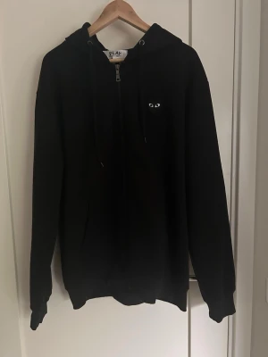 Svart hoodie från Comme des Garçons Play - Svart hoodie med dragkedja från Comme des Garçons Play. Har huva med dragsko och ikoniskt hjärta med ögon-tryck på ryggen samt mindre logga på bröstet. Mjuk bomullskvalitet och ribbade muddar vid ärmslut och nederkant. Tröjan passar som L  även fast det står på tröjan 3 xxl 
