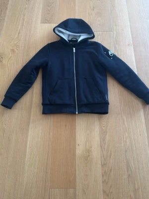 Svart hoodie med dragkedja och huva från moose knuckels  - Snygg svart hoodie från moose knuckels med dragkedja framtill och två stora fickor. Utrustad med huva som har ljus insida och en cool logga på ena ärmen. Perfekt för en chill och avslappnad stil. Det är storlek XL barn passar 164-170