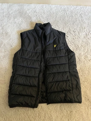 Svart dunväst från Lyle & Scott - Stilren svart dunväst från Lyle & Scott med gul logga på bröstet. Västen har hög krage, quiltad design och dragkedja framtill. Perfekt att slänga över en hoodie för extra värme. Materialet är glansigt och lätt vadderat.