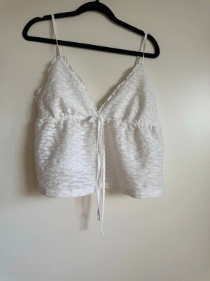 Vit linnetopp med knyt och struktur - Snygg vit linnetopp med smala axelband och v-ringning. Toppen har en strukturerad yta och knytning framtill som ger en söt och trendig look. Perfekt till jeans eller kjol för en fräsch stil.