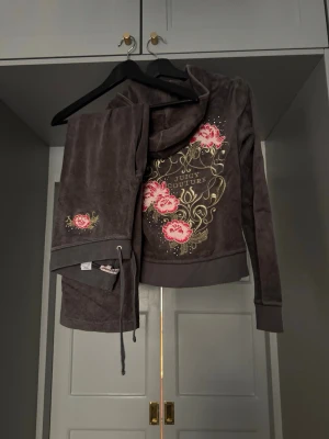 Juicy Couture grå velourset - Mysigt loungewear-set i grå velour från Juicy Couture. Setet består av en zip-hoodie med guldiga detaljer, rosa blommor och logga på ryggen samt matchande byxor med broderad blomdetalj. Perfekt för chill dagar hemma.