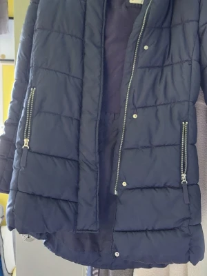 Mörkblå pufferjacka med huva - Snygg mörkblå pufferjacka med huva och dragkedjor på sidorna. Jackan har quiltad design, två fickor med dragkedja och stängs med både dragkedja och tryckknappar. Perfekt för kalla vinterdagar och riktigt skön att bära.