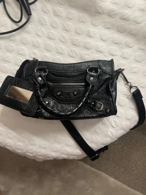 Svart handväska - Cool svart handväska med silverfärgade detaljer, dragkedja framtill och dekorativa nitar. Väskan har både handtag och avtagbar axelrem samt spegelaccessoar. Perfekt nu till sommaren och för dig som gillar edgy och tidlös design. Har köpt second hand för 1500kr men bara använt fåtal gånger själv. Priset kan diskuteras!💓  #handväska #svarthandväska #citybag