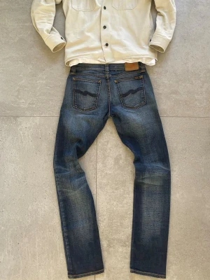 Blå jeans från Nudie Jeans - Storlek W29 L32, modell : Tight long John, Snygga blå jeans från Nudie Jeans med klassisk femficksdesign och tydliga slitningar. Jeansen har raka ben och ikoniska broderade bakfickor. Tillverkade i denim med en autentisk tvättad look och läderpatch bak i midjan. Tveka inte att skriva vid funderingar!!!