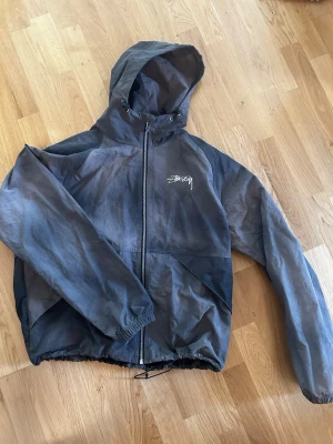 Svart vindjacka från Stüssy - Svart/grå vindjacka från Stüssy med vit logga på bröstet och stor logga på ryggen. Jackan har huva med dragsko, hel dragkedja framtill och elastiska muddar. Materialet är tunt och lätt, perfekt för blåsiga dagar. 