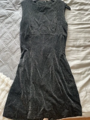 Svart glittrig kort klänning - Snygg svart kort klänning med glittrigt tyg och ärmlös design. Klänningen har en figurnära passform och rund halsringning, perfekt för att sticka ut. Materialet känns mjukt och följsamt mot kroppen.