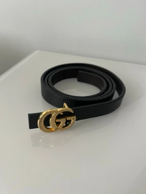 Svart bälte från Gucci med GG-spänne - Snyggt svart bälte från Gucci med klassiskt dubbelt GG-spänne i guld. Bältet är smalt och gjort i skinn, perfekt för att lyfta vilken outfit som helst med en lyxig touch. Lite repad. Se bild. 