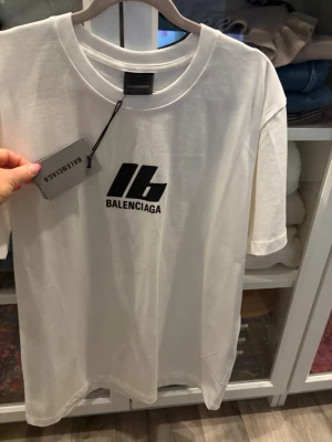 Vit Balenciaga t-shirt med tryck - Vit t-shirt från Balenciaga med svart logotyptryck framtill och stor text på ryggen. Klassisk rund halsringning och korta ärmar. Tillverkad i mjuk bomull för en skön och avslappnad känsla.