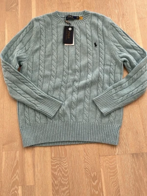 Ljusblå kabelstickad tröja Polo Ralph Lauren - Kabelstickad ljusblå tröja från Polo Ralph Lauren med rund halsringning och broderad logga på bröstet. Tröjan har ribbade muddar vid ärmslut och nederkant, och är tillverkad i mjukt material som passar perfekt till en avslappnad men stilren look.