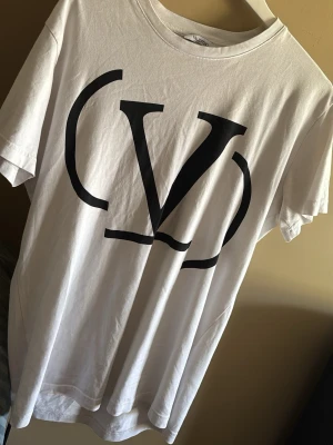 Vit Valentino t-shirt med stor logga - Vit t-shirt från Valentino med stor svart logga framtill. Klassisk rund halsringning och korta ärmar. Tillverkad i mjuk bomull för en skön och avslappnad känsla. Perfekt för dig som gillar stilrena och trendiga plagg.