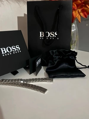 Silverfärgat armband från Hugo Boss - Stilrent armband i silverfärgat rostfritt stål från Hugo Boss. Armbandet har en länkdesign med gravering av BOSS på spännet. Perfekt accessoar för en modern look och levereras med originalpåse och presentförpackning.