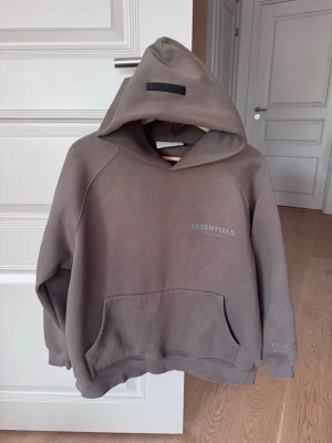 Brun Essentials hoodie med huva - Snygg brun hoodie från Essentials med huva och stor magficka. Diskret logga på bröstet och ärmen. Perfekt för en chill och avslappnad stil. Färg: Taupe
