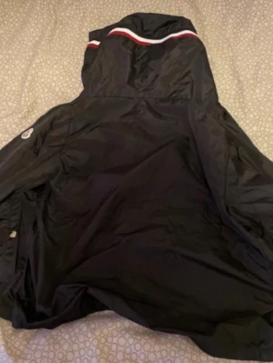 Svart Moncler vindjacka med huva - Svart vindjacka från Moncler med huva och vit-röd randdetalj upptill. Jackan har Moncler-logga på ärmen och är tillverkad i ett lätt, vindtätt material. Perfekt för blåsiga dagar och snygg streetstil.