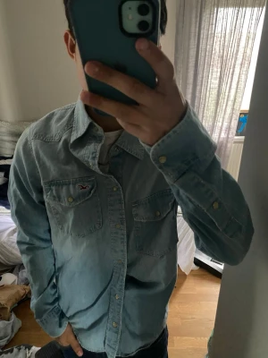 Ljusblå jeansskjorta från Hollister - Säljer en snygg ljusblå jeansskjorta från Hollister med klassisk krage och två bröstfickor med knappar. Skjortan har långa ärmar och är tillverkad i bomull. Perfekt för en avslappnad och cool stil.