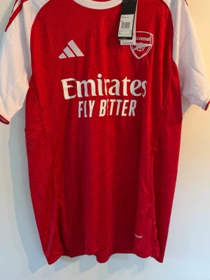 Arsenal matchtröja Gyökeres 25/26 - Röd och vit Arsenal matchtröja från Adidas med Gyökeres och nummer 14 på ryggen. Tröjan har klubbmärke och Adidas-logga på bröstet samt Emirates Fly Better-tryck. Tillverkad i lätt och ventilerande material, perfekt för fotbollsträning eller match.