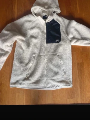 Vit teddy-hoodie från Nike - Mysig vit hoodie från Nike i teddyfleece med dragkedjor på fickorna och en svart bröstficka med Nike-logga. Hoodien har luva och en avslappnad passform, perfekt för chill dagar. Materialet är fluffigt och ger en cool streetwear-vibe.