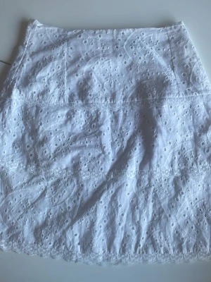 Vit broderad kjol från Escape - Superfin vit kortkjol från Escape med broderade blommönster och diskreta spetsdetaljer. Kjolen har dragkedja i bak och är gjord i bomull, perfekt för en fräsch och somrig look. 