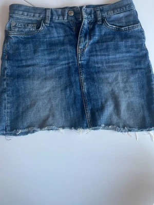 Blå jeanskjol med rå kant - Snygg blå jeanskjol i kort modell med fransig kant nertill. Klassisk femficksdesign med två bakfickor med lock och knappar. Kjolen har bälteshällor. Perfekt för dig som gillar en avslappnad och trendig stil.