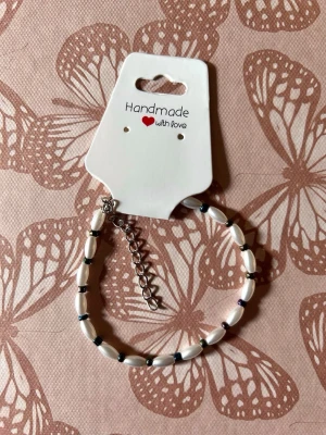 Handgjort pärlarmband med vita pärlor - Ett handgjort armband med avlånga vita pärlor och små svarta detaljer. Armbandet har en justerbar silverfärgad kedja och passar perfekt för dig som gillar unika accessoarer. Stilren och enkel design som lyfter din look.