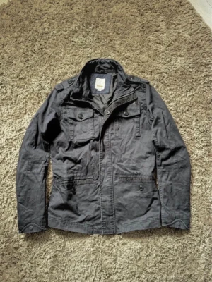 militärjacka från Diesel - Snygg svart militärjacka från Diesel med fyra framfickor, axelklaffar och dragkedja. Jackan har en klassisk militärinspirerad look och är tillverkad i slitstark bomull. Perfekt för dig som gillar streetstyle och vill ha en stilren jacka med coola detaljer. Den är M men skulle kunna passa en lite större S också 