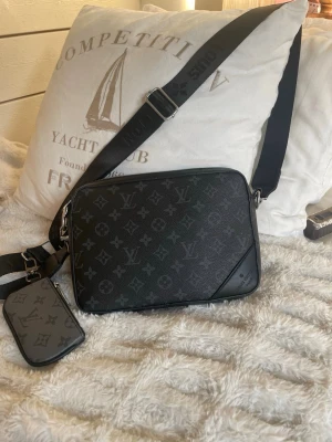 Louis Vuitton svart axelväska monogram - Snygg svart axelväska från Louis Vuitton med klassiskt monogrammönster i grått och svart. Väskan har justerbar axelrem och en matchande liten pouch som hänger på remmen. Tillverkad i tåligt lädermaterial med silverfärgade detaljer.