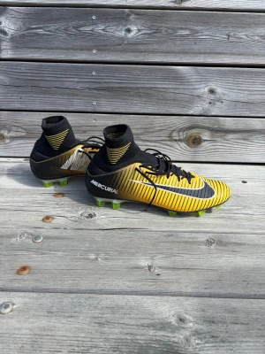 Nike Mercurial gula fotbollsskor - Säljer ett par Nike Mercurial fotbollsskor i svart och gult med coola ränder och högt skaft. Skorna har snörning, rund tå och gröna dobbar för bra grepp på planen. Perfekta för dig som vill sticka ut på fotbollsplanen.