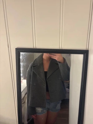 Grå skepparkavaj med bred krage - Stilren grå skepparkavaj med dubbelknäppning och bred krage. Jackan är kortare i modellen och har en klassisk, rak passform. Perfekt för dig som vill ha en tidlös look med lite edge. Materialet ser ut att vara ullblandning.