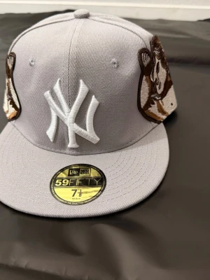 Grå New Era Yankees keps 59FIFTY - Snygg grå 59FIFTY keps från New Era med New York Yankees-logga framtill och broderade motiv på sidorna. Baksidan har texten 'Com Stan' i vit brodyr. Platt skärm och klassisk passform, tillverkad i bomull.