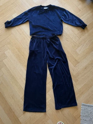 Mörkblått velourset loungewear - Mysigt tvådelat loungewear-set i mörkblå velour. Setet består av en långärmad tröja med rund hals och ett par vida byxor med resår i midjan. Perfekt för chill dagar hemma. Materialet är mjukt och glansigt för extra komfort.