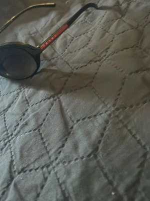 Svarta solglasögon från Prada - Snygga svarta solglasögon från Prada med rundad form och mörka glas. Bågarna har en röd detalj med Prada-loggan på sidan. Perfekta för att lyfta din stil och ge en exklusiv känsla till din outfit.