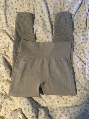 Grå sömlösa leggings med hög midja - Gråa sömlösa leggings med hög midja och ribbade detaljer. Baksidan har scrunch-effekt och markerade partier för en snygg passform. Mjukt och stretchigt material som sitter skönt på kroppen. Perfekta för träning eller chill.